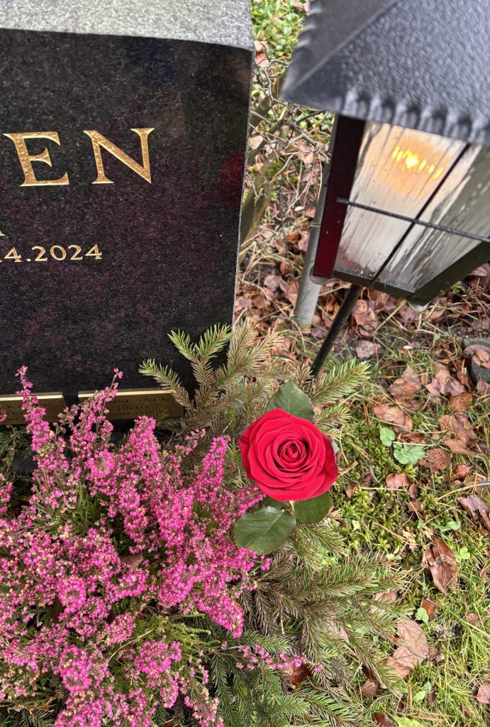 Osa hautakivestä näkyy, sen edessä kanervia, kynttilälyhyty ja ruusu